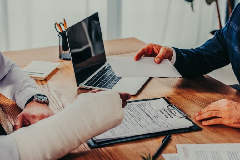 cropped view of businessman sitting at table and giving paper sheet to worker with broken arm in office, compensation concept